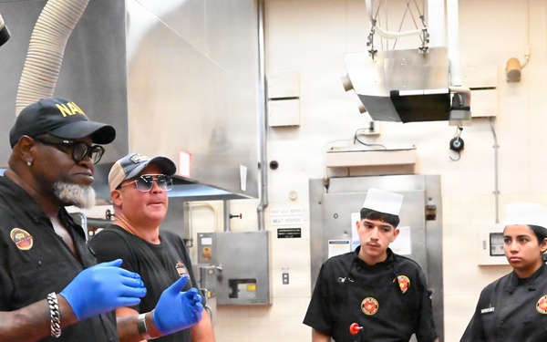 A culinary instruction session is taking place in a professional kitchen, where two instructors wearing black shirts and blue latex gloves appear to be demonstrating cooking techniques to three students. The students are dressed in black chef uniforms with matching patches/logos, and one wears a traditional white chef's toque. A red cutting board with meat and various condiment bottles and sauces are visible on the work table.