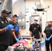 A culinary instruction session is taking place in a professional kitchen, where two instructors wearing black shirts and blue latex gloves appear to be demonstrating cooking techniques to three students. The students are dressed in black chef uniforms with matching patches/logos, and one wears a traditional white chef's toque. A red cutting board with meat and various condiment bottles and sauces are visible on the work table.