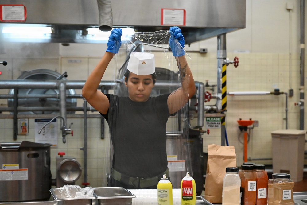 A kitchen worker wearing blue latex gloves and a white chef's hat is stretching or unrolling clear plastic wrap in what appears to be a commercial or institutional kitchen. The worker is dressed in a black t-shirt and is standing at a stainless steel prep table with various cooking supplies visible in the foreground. The setting appears to be a military or institutional food service facility based on the signage and equipment visible.