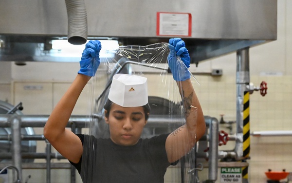 A kitchen worker wearing blue latex gloves and a white chef's hat is stretching or unrolling clear plastic wrap in what appears to be a commercial or institutional kitchen. The worker is dressed in a black t-shirt and is standing at a stainless steel prep table with various cooking supplies visible in the foreground. The setting appears to be a military or institutional food service facility based on the signage and equipment visible.