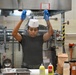 A kitchen worker wearing blue latex gloves and a white chef's hat is stretching or unrolling clear plastic wrap in what appears to be a commercial or institutional kitchen. The worker is dressed in a black t-shirt and is standing at a stainless steel prep table with various cooking supplies visible in the foreground. The setting appears to be a military or institutional food service facility based on the signage and equipment visible.