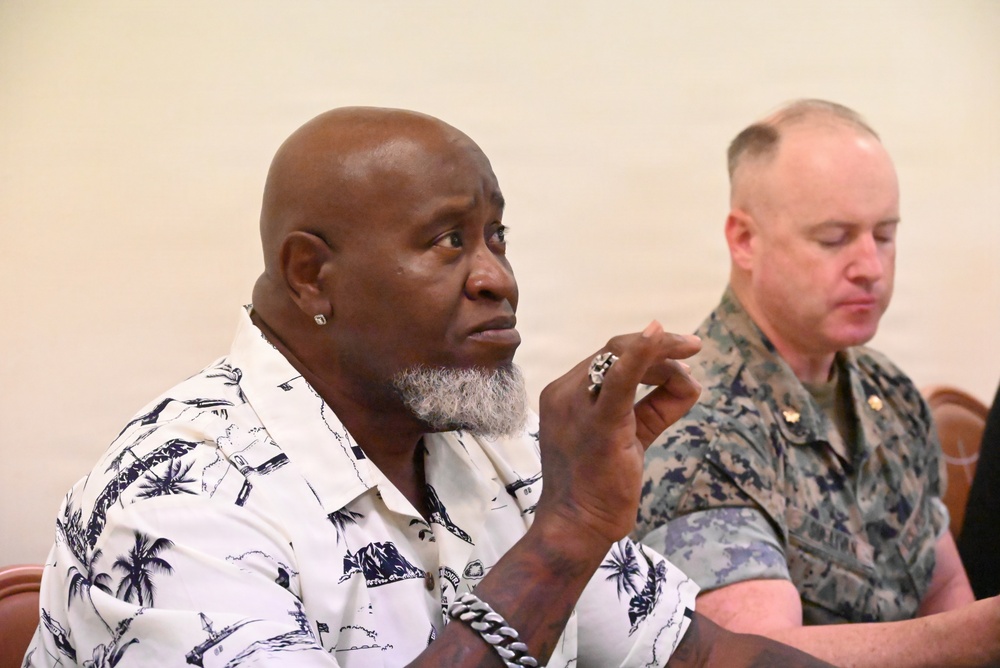 Two men are seated in what appears to be a formal meeting or briefing setting. The man in the foreground is wearing a tropical-print shirt and has a gray beard, appearing attentive with his hand raised near his chin. Behind him sits a man in U.S. Marine Corps camouflage uniform who appears to be looking downward.