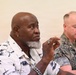 Two men are seated in what appears to be a formal meeting or briefing setting. The man in the foreground is wearing a tropical-print shirt and has a gray beard, appearing attentive with his hand raised near his chin. Behind him sits a man in U.S. Marine Corps camouflage uniform who appears to be looking downward.