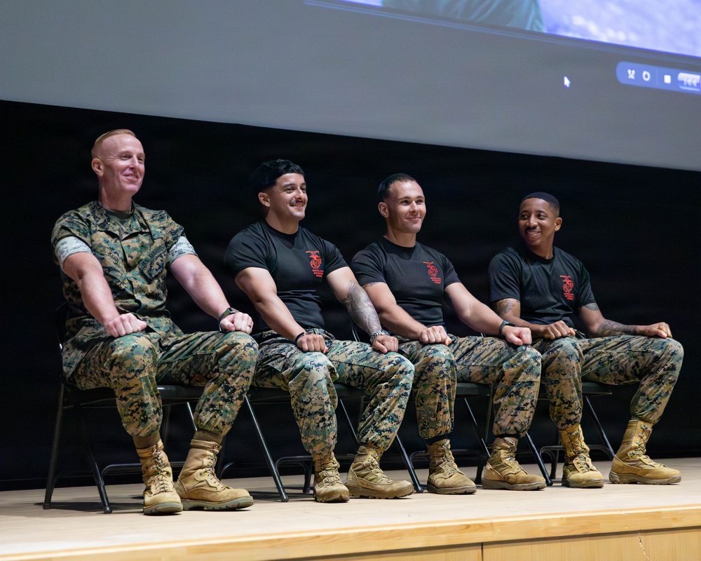 Four U.S. Marines are seated on a stage in a row on folding chairs, appearing to be participating in a panel discussion or presentation event. One Marine on the left wears a full camouflage utility uniform, while the other three wear black t-shirts with a red Marine Corps emblem and camouflage trousers. All four are smiling and appear relaxed, suggesting an informal or community engagement setting.
