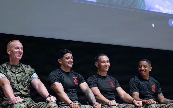 Four U.S. Marines are seated on a stage in a row on folding chairs, appearing to be participating in a panel discussion or presentation event. One Marine on the left wears a full camouflage utility uniform, while the other three wear black t-shirts with a red Marine Corps emblem and camouflage trousers. All four are smiling and appear relaxed, suggesting an informal or community engagement setting.