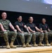 Four U.S. Marines are seated on a stage in a row on folding chairs, appearing to be participating in a panel discussion or presentation event. One Marine on the left wears a full camouflage utility uniform, while the other three wear black t-shirts with a red Marine Corps emblem and camouflage trousers. All four are smiling and appear relaxed, suggesting an informal or community engagement setting.