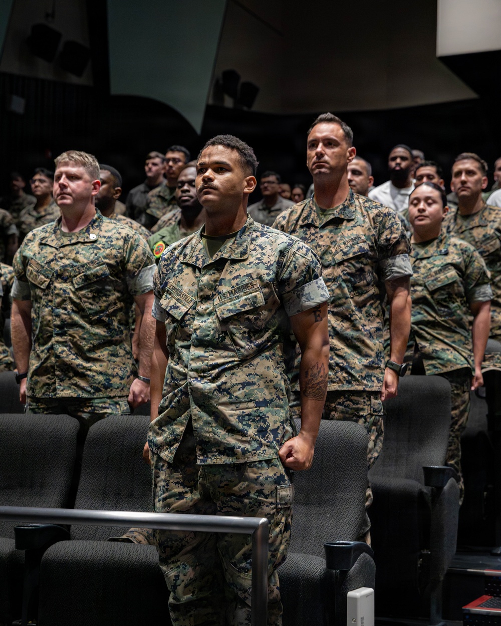 A large group of U.S. Marines in MARPAT woodland digital camouflage uniforms are standing at attention in what appears to be an auditorium or theater setting. The Marines are arranged in rows among theater-style seating, suggesting a formal military ceremony, briefing, or formation event. The foreground Marine's name tape and 'U.S. Marines' branch tape are clearly visible.