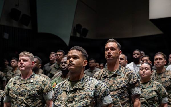 A large group of U.S. Marines in MARPAT woodland digital camouflage uniforms are standing at attention in what appears to be an auditorium or theater setting. The Marines are arranged in rows among theater-style seating, suggesting a formal military ceremony, briefing, or formation event. The foreground Marine's name tape and 'U.S. Marines' branch tape are clearly visible.