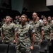 A large group of U.S. Marines in MARPAT woodland digital camouflage uniforms are standing at attention in what appears to be an auditorium or theater setting. The Marines are arranged in rows among theater-style seating, suggesting a formal military ceremony, briefing, or formation event. The foreground Marine's name tape and 'U.S. Marines' branch tape are clearly visible.