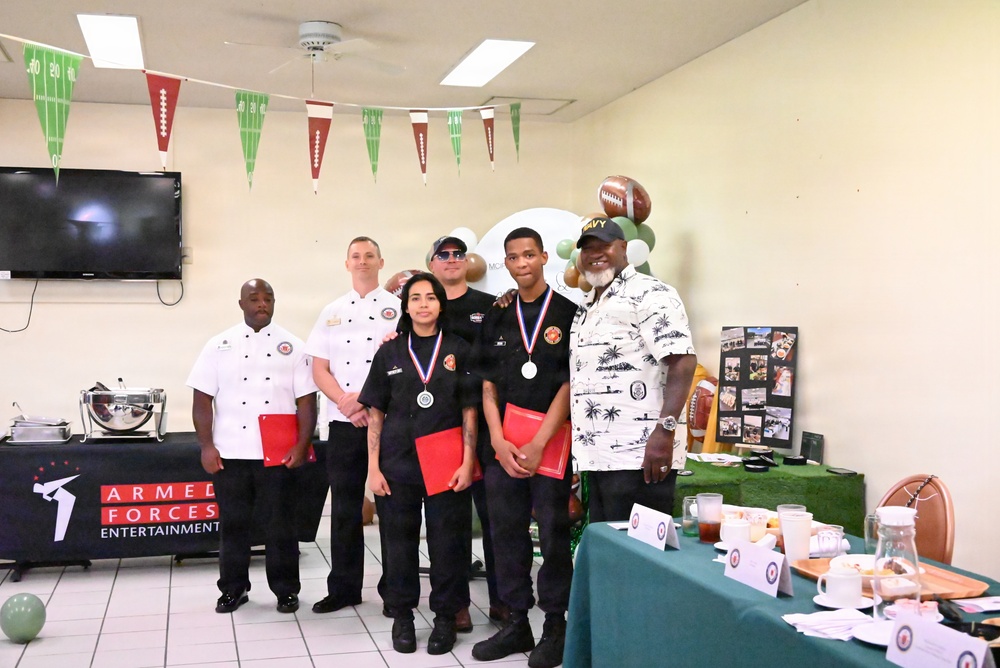 A group of six people pose for a photo at what appears to be an Armed Forces Entertainment event with a football/sports theme. Three individuals in the center are wearing medals and holding red folders, suggesting they are award recipients. The setting appears to be an indoor dining or event hall decorated with football-themed bunting and balloons.
