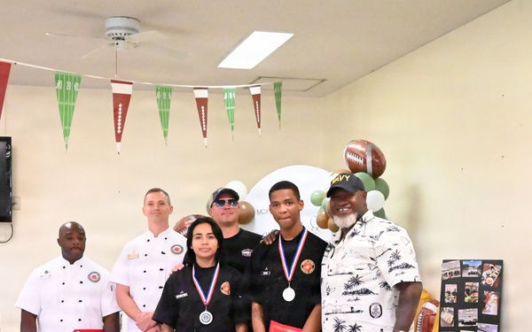A group of six people pose for a photo at what appears to be an Armed Forces Entertainment event with a football/sports theme. Three individuals in the center are wearing medals and holding red folders, suggesting they are award recipients. The setting appears to be an indoor dining or event hall decorated with football-themed bunting and balloons.
