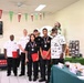 A group of six people pose for a photo at what appears to be an Armed Forces Entertainment event with a football/sports theme. Three individuals in the center are wearing medals and holding red folders, suggesting they are award recipients. The setting appears to be an indoor dining or event hall decorated with football-themed bunting and balloons.