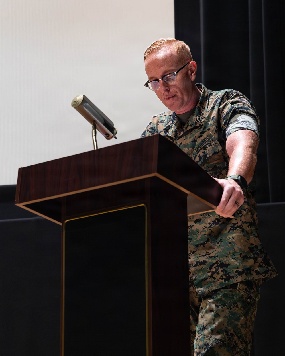 A U.S. Marine in MARPAT woodland digital camouflage uniform stands at a wooden podium, appearing to be delivering a speech or presentation. The individual wears glasses and has short reddish hair, with rank insignia and a name tape visible on the uniform. The setting appears to be an indoor auditorium or formal briefing room.
