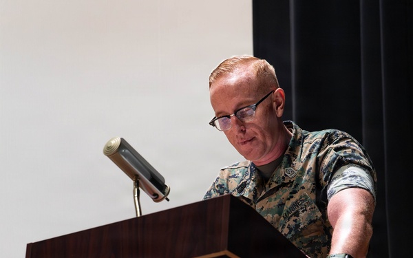 A U.S. Marine in MARPAT woodland digital camouflage uniform stands at a wooden podium, appearing to be delivering a speech or presentation. The individual wears glasses and has short reddish hair, with rank insignia and a name tape visible on the uniform. The setting appears to be an indoor auditorium or formal briefing room.