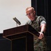 A U.S. Marine in MARPAT woodland digital camouflage uniform stands at a wooden podium, appearing to be delivering a speech or presentation. The individual wears glasses and has short reddish hair, with rank insignia and a name tape visible on the uniform. The setting appears to be an indoor auditorium or formal briefing room.