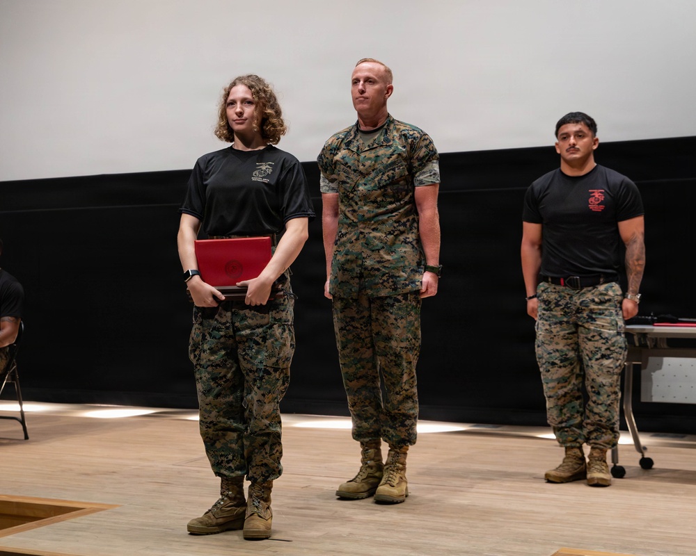 Three military personnel, appearing to be U.S. Marines based on their MARPAT camouflage uniforms and black PT shirts with Marine Corps insignia, are standing on a stage or platform. A female Marine on the left is holding a red presentation folder or award case, suggesting a recognition or awards ceremony is taking place. A senior Marine in full camouflage uniform stands beside her, while a third Marine in a black PT shirt stands at ease in the background.