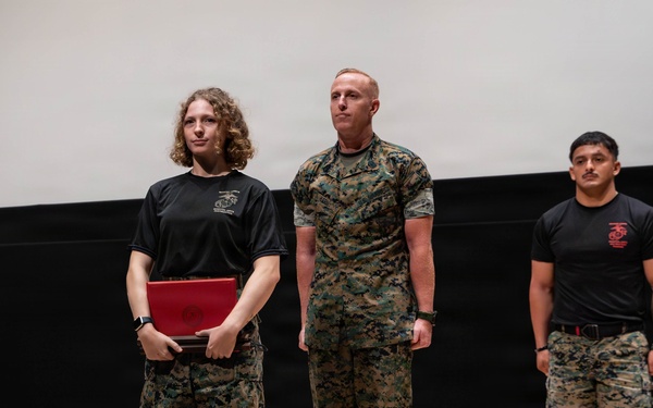 Three military personnel, appearing to be U.S. Marines based on their MARPAT camouflage uniforms and black PT shirts with Marine Corps insignia, are standing on a stage or platform. A female Marine on the left is holding a red presentation folder or award case, suggesting a recognition or awards ceremony is taking place. A senior Marine in full camouflage uniform stands beside her, while a third Marine in a black PT shirt stands at ease in the background.
