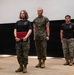 Three military personnel, appearing to be U.S. Marines based on their MARPAT camouflage uniforms and black PT shirts with Marine Corps insignia, are standing on a stage or platform. A female Marine on the left is holding a red presentation folder or award case, suggesting a recognition or awards ceremony is taking place. A senior Marine in full camouflage uniform stands beside her, while a third Marine in a black PT shirt stands at ease in the background.