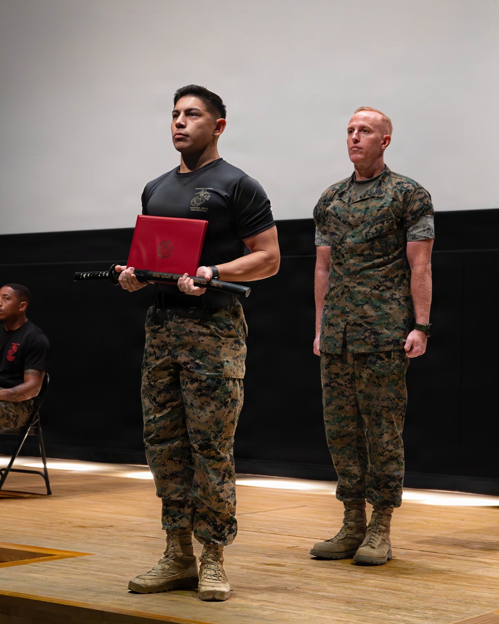 Two U.S. Marines are standing on a wooden stage during what appears to be a formal ceremony. The Marine in the foreground is wearing a black 'Marine Corps Martial Arts Instructor' t-shirt and camouflage trousers, holding a red certificate folder and a black ceremonial sword (katana-style). A second Marine in full MARPAT camouflage uniform stands at attention in the background.