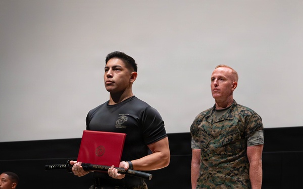 Two U.S. Marines are standing on a wooden stage during what appears to be a formal ceremony. The Marine in the foreground is wearing a black 'Marine Corps Martial Arts Instructor' t-shirt and camouflage trousers, holding a red certificate folder and a black ceremonial sword (katana-style). A second Marine in full MARPAT camouflage uniform stands at attention in the background.