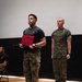 Two U.S. Marines are standing on a wooden stage during what appears to be a formal ceremony. The Marine in the foreground is wearing a black 'Marine Corps Martial Arts Instructor' t-shirt and camouflage trousers, holding a red certificate folder and a black ceremonial sword (katana-style). A second Marine in full MARPAT camouflage uniform stands at attention in the background.