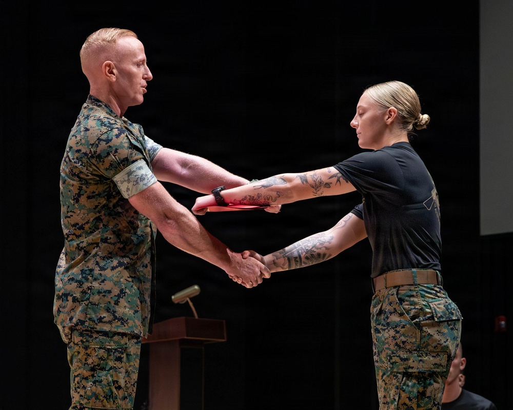 A military ceremony is taking place in which a male service member in full camouflage uniform is presenting a red folder or certificate to a female service member while shaking hands. Both individuals are wearing U.S. Marine Corps MARPAT (digital camouflage) uniforms. The setting appears to be an indoor auditorium or ceremonial hall with a podium visible in the background.