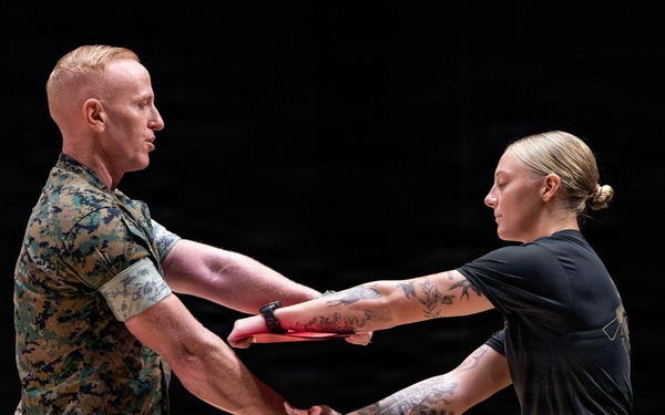 A military ceremony is taking place in which a male service member in full camouflage uniform is presenting a red folder or certificate to a female service member while shaking hands. Both individuals are wearing U.S. Marine Corps MARPAT (digital camouflage) uniforms. The setting appears to be an indoor auditorium or ceremonial hall with a podium visible in the background.