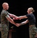 A military ceremony is taking place in which a male service member in full camouflage uniform is presenting a red folder or certificate to a female service member while shaking hands. Both individuals are wearing U.S. Marine Corps MARPAT (digital camouflage) uniforms. The setting appears to be an indoor auditorium or ceremonial hall with a podium visible in the background.