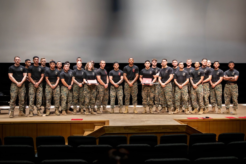 A large group of military personnel, approximately 25-30 individuals, are posed in a single row on an auditorium or theater stage. They are wearing matching black t-shirts with unit insignia and camouflage (MARPAT) trousers with tan boots, consistent with U.S. Marine Corps uniforms. Several individuals in the group are holding red folders or certificates, suggesting a formal recognition or graduation ceremony.
