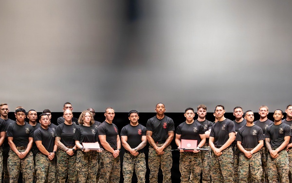 A large group of military personnel, approximately 25-30 individuals, are posed in a single row on an auditorium or theater stage. They are wearing matching black t-shirts with unit insignia and camouflage (MARPAT) trousers with tan boots, consistent with U.S. Marine Corps uniforms. Several individuals in the group are holding red folders or certificates, suggesting a formal recognition or graduation ceremony.