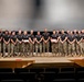 A large group of military personnel, approximately 25-30 individuals, are posed in a single row on an auditorium or theater stage. They are wearing matching black t-shirts with unit insignia and camouflage (MARPAT) trousers with tan boots, consistent with U.S. Marine Corps uniforms. Several individuals in the group are holding red folders or certificates, suggesting a formal recognition or graduation ceremony.