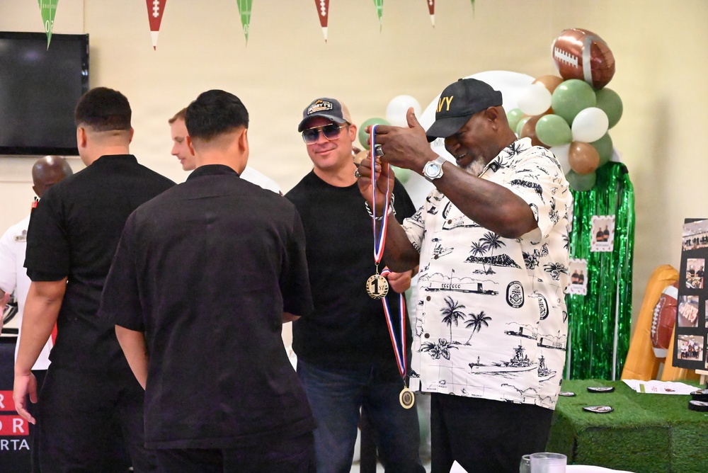 A group of people are gathered at what appears to be a football-themed event or ceremony, likely a military or community function. A man wearing a Navy cap and Hawaiian shirt is presenting or displaying a gold medal on a red, white, and blue ribbon to another individual. The setting is decorated with football-themed balloons, pennants, and green tinsel curtains.