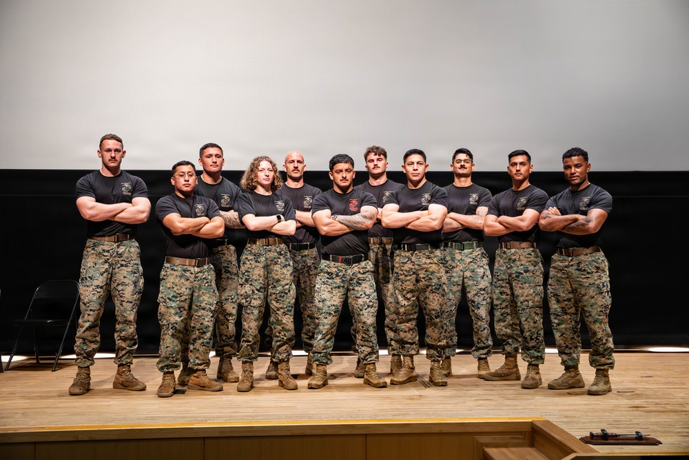 A group of twelve military personnel, identifiable as U.S. Marines based on their uniforms, pose together on a stage or platform. All individuals are wearing black t-shirts with Marine Corps insignia and digital camouflage (MARPAT) trousers with tan combat boots. They are standing in a unified pose with arms crossed, suggesting a formal team or unit photograph.