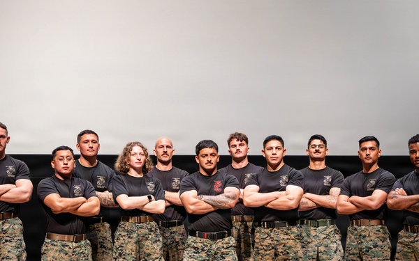 A group of twelve military personnel, identifiable as U.S. Marines based on their uniforms, pose together on a stage or platform. All individuals are wearing black t-shirts with Marine Corps insignia and digital camouflage (MARPAT) trousers with tan combat boots. They are standing in a unified pose with arms crossed, suggesting a formal team or unit photograph.