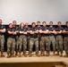 A group of twelve military personnel, identifiable as U.S. Marines based on their uniforms, pose together on a stage or platform. All individuals are wearing black t-shirts with Marine Corps insignia and digital camouflage (MARPAT) trousers with tan combat boots. They are standing in a unified pose with arms crossed, suggesting a formal team or unit photograph.