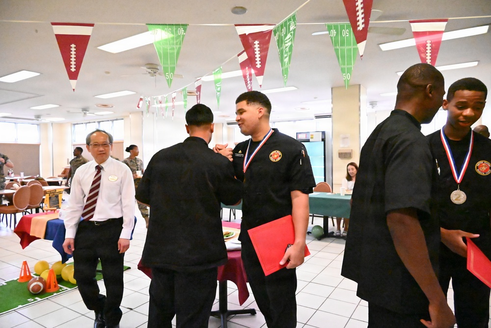 A medal ceremony is taking place in a decorated dining facility or cafeteria, where individuals in black uniforms are receiving medals and red folders. The room is decorated with football-themed pennant banners in red and green, suggesting a sports-themed event. An older gentleman in a white shirt and red striped tie appears to be an official or judge observing the proceedings.