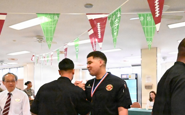 A medal ceremony is taking place in a decorated dining facility or cafeteria, where individuals in black uniforms are receiving medals and red folders. The room is decorated with football-themed pennant banners in red and green, suggesting a sports-themed event. An older gentleman in a white shirt and red striped tie appears to be an official or judge observing the proceedings.