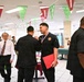 A medal ceremony is taking place in a decorated dining facility or cafeteria, where individuals in black uniforms are receiving medals and red folders. The room is decorated with football-themed pennant banners in red and green, suggesting a sports-themed event. An older gentleman in a white shirt and red striped tie appears to be an official or judge observing the proceedings.