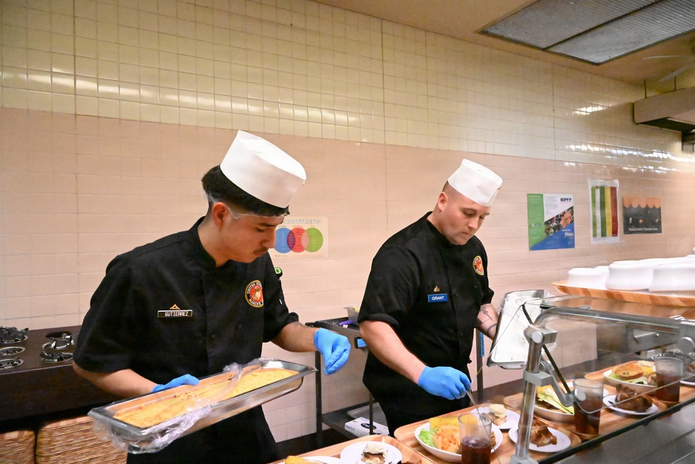 Two food service workers wearing black uniforms, white chef hats, and blue latex gloves are working at a serving line or buffet station. Their name tags identify them as 'GUTIERREZ' (left) and 'GRANT' (right), and both wear patches consistent with military or institutional food service. They appear to be preparing and plating food items at a cafeteria-style counter.