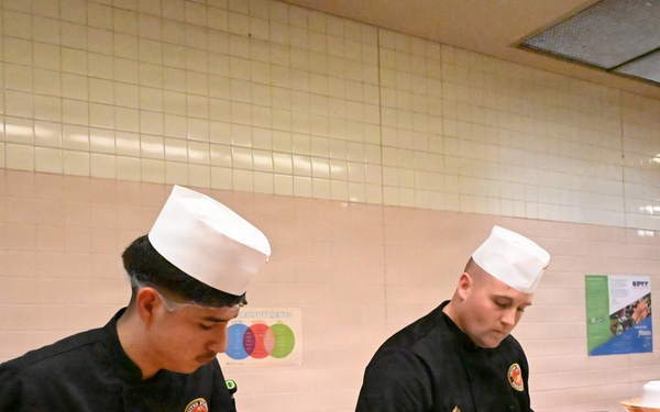 Two food service workers wearing black uniforms, white chef hats, and blue latex gloves are working at a serving line or buffet station. Their name tags identify them as 'GUTIERREZ' (left) and 'GRANT' (right), and both wear patches consistent with military or institutional food service. They appear to be preparing and plating food items at a cafeteria-style counter.
