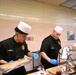 Two food service workers wearing black uniforms, white chef hats, and blue latex gloves are working at a serving line or buffet station. Their name tags identify them as 'GUTIERREZ' (left) and 'GRANT' (right), and both wear patches consistent with military or institutional food service. They appear to be preparing and plating food items at a cafeteria-style counter.
