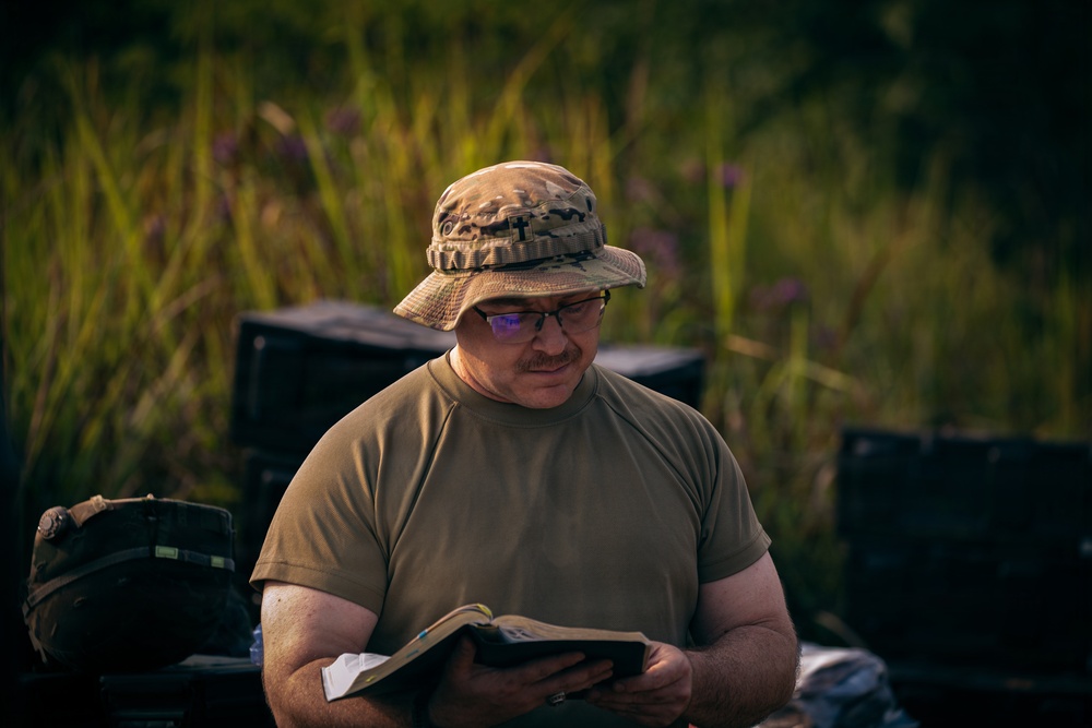 A middle-aged man wearing military-style olive drab clothing and a camouflage boonie hat is reading a book outdoors. He is wearing glasses and appears focused on the text. The setting appears to be a field or natural area with tall green grass and purple wildflowers in the background.