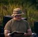 A middle-aged man wearing military-style olive drab clothing and a camouflage boonie hat is reading a book outdoors. He is wearing glasses and appears focused on the text. The setting appears to be a field or natural area with tall green grass and purple wildflowers in the background.