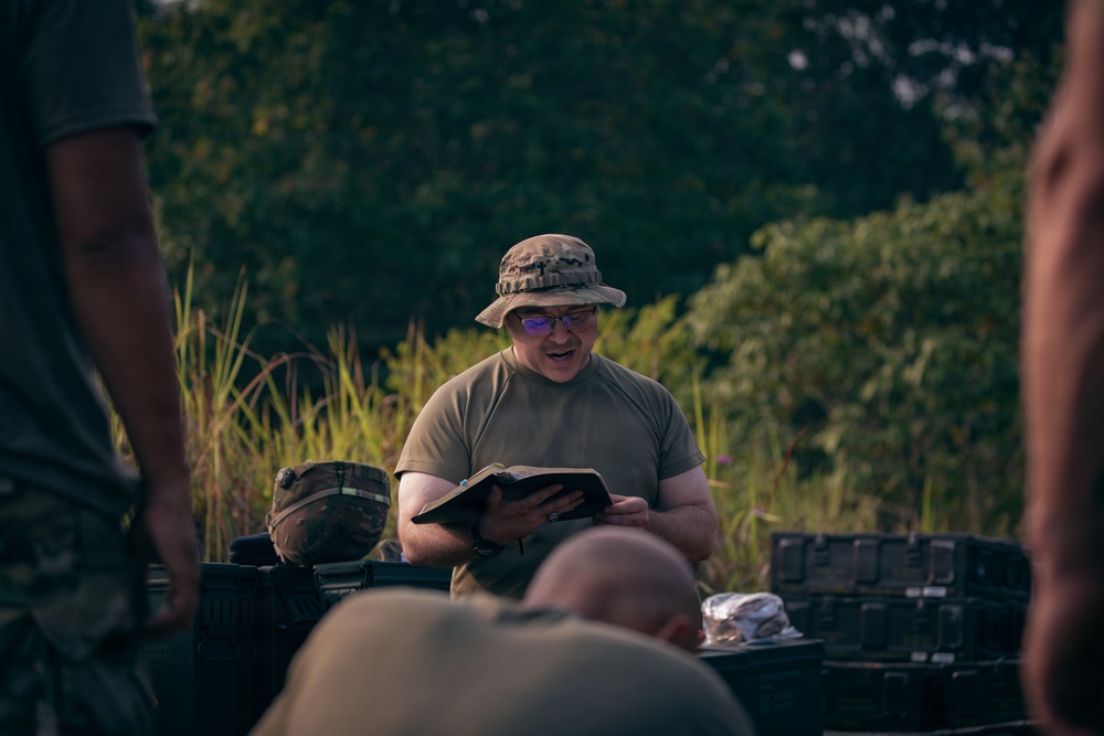 A person in military-style clothing and a camouflage boonie hat is reading aloud from a book, likely a Bible or field manual, to a small group gathered outdoors. The setting appears to be a field or wooded area with military equipment visible in the background. The scene suggests an informal gathering, possibly a field religious service or briefing.