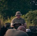 A person in military-style clothing and a camouflage boonie hat is reading aloud from a book, likely a Bible or field manual, to a small group gathered outdoors. The setting appears to be a field or wooded area with military equipment visible in the background. The scene suggests an informal gathering, possibly a field religious service or briefing.