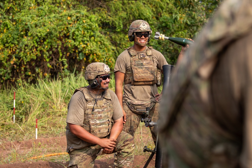 Two military personnel in full combat gear, including helmets, ballistic vests, and sunglasses, are positioned near what appears to be a mortar system in an outdoor training environment. Both soldiers are smiling, suggesting a training exercise rather than a combat situation. A third partially visible individual in camouflage is present in the foreground right.