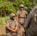 Two military personnel in full combat gear, including helmets, ballistic vests, and sunglasses, are positioned near what appears to be a mortar system in an outdoor training environment. Both soldiers are smiling, suggesting a training exercise rather than a combat situation. A third partially visible individual in camouflage is present in the foreground right.