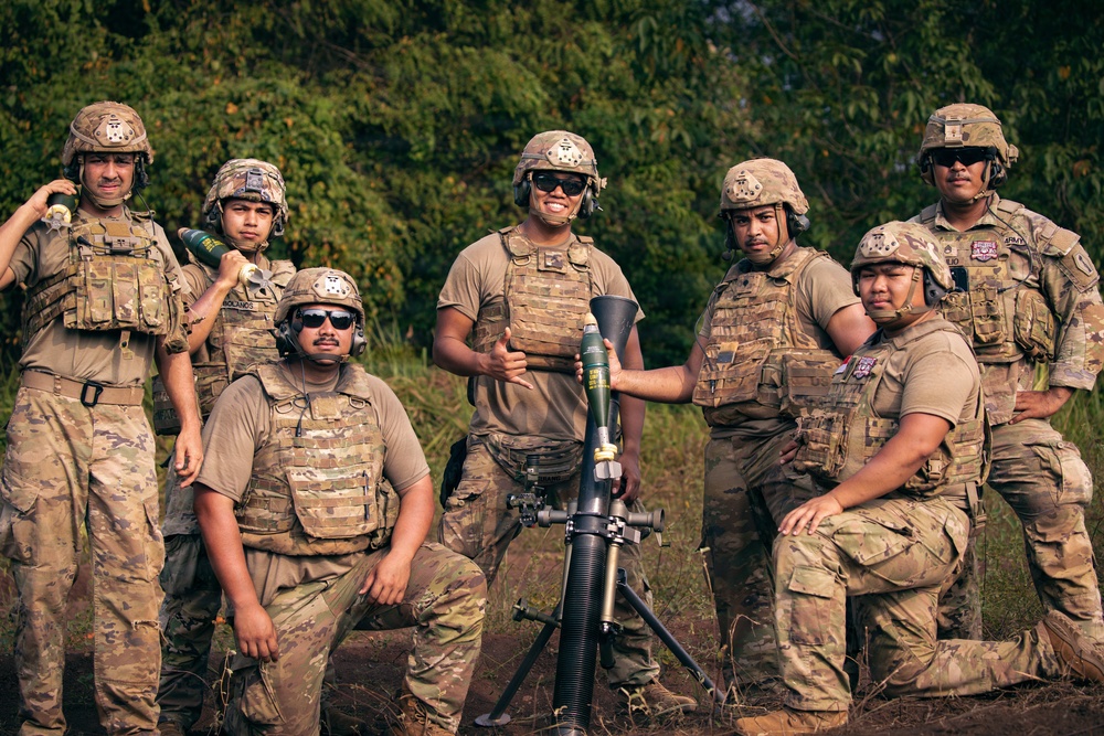 A group of seven military personnel in U.S. Army uniforms pose together outdoors with a mortar system and a mortar round. They are wearing multicam camouflage uniforms, plate carrier vests, and combat helmets, suggesting a training exercise or field operation. The group appears relaxed and is posing for a group photograph in a wooded outdoor setting.
