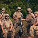 A group of seven military personnel in U.S. Army uniforms pose together outdoors with a mortar system and a mortar round. They are wearing multicam camouflage uniforms, plate carrier vests, and combat helmets, suggesting a training exercise or field operation. The group appears relaxed and is posing for a group photograph in a wooded outdoor setting.