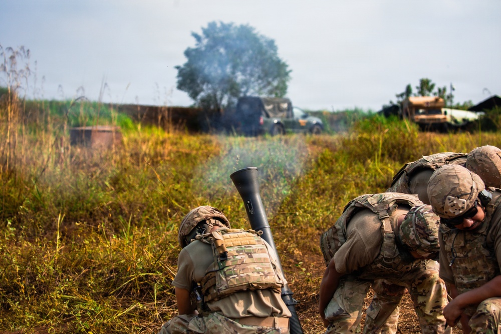 Military personnel in camouflage uniforms and tactical gear are operating a mortar system in an open field environment. Smoke is visible rising from the mortar tube, indicating a recent or ongoing firing sequence. Military vehicles are visible in the background amid tall grass and trees.